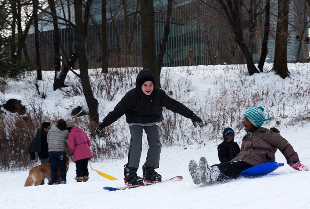 A boy slides down the snow-covered hill on a snowboard next to a girl on blue sled.