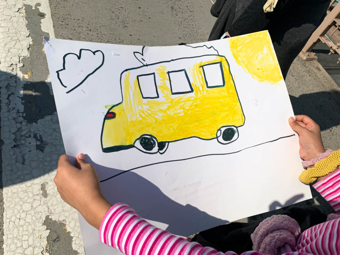 During the march over the Brooklyn Bridge, a child holds a drawing of a short yellow bus, usually used for transporting students with special needs. Mar 19, 2022. (Photo by Eleonora Francica for NY City Lens)